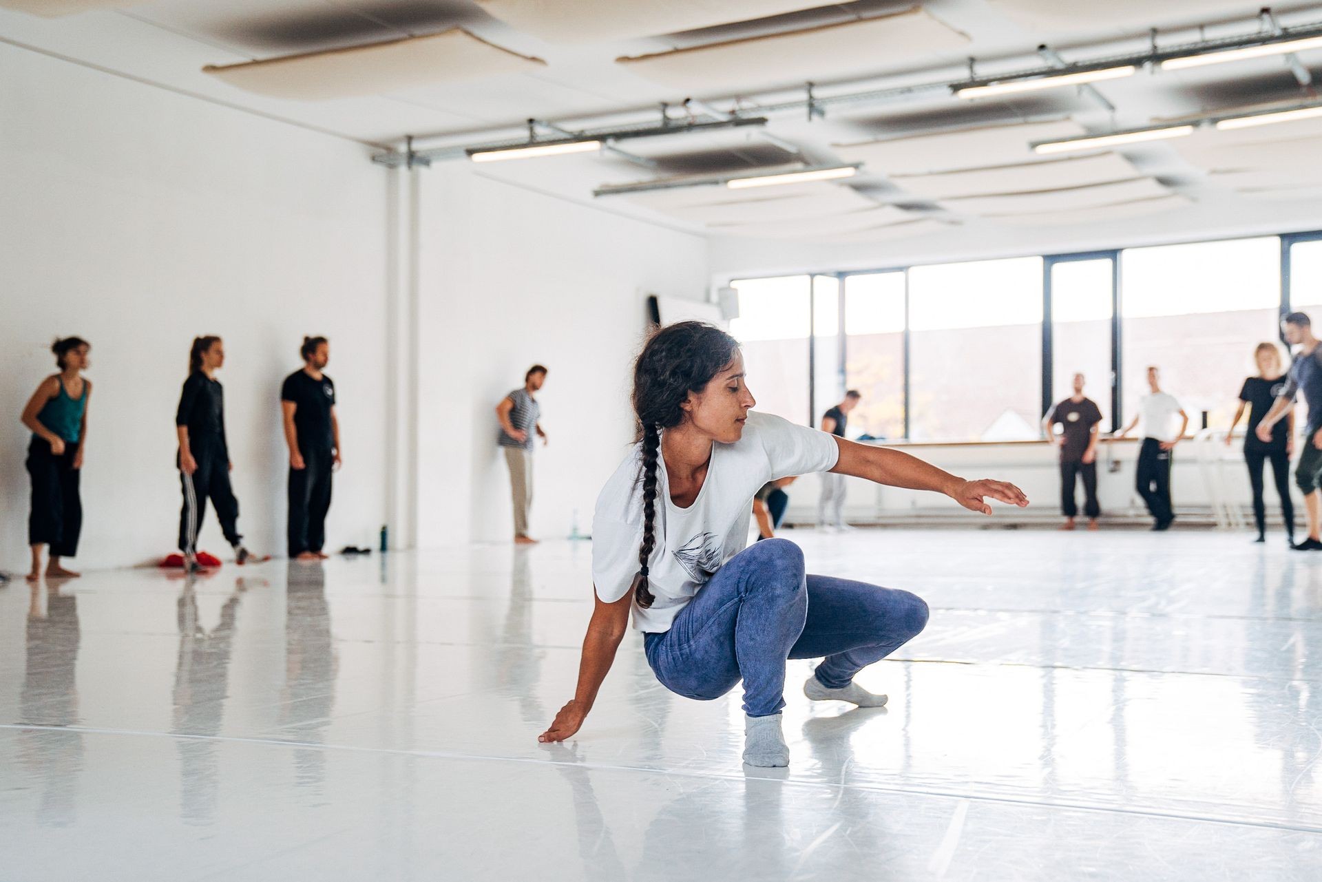 Dancer practicing movement in a studio while others observe, with large windows in the background.