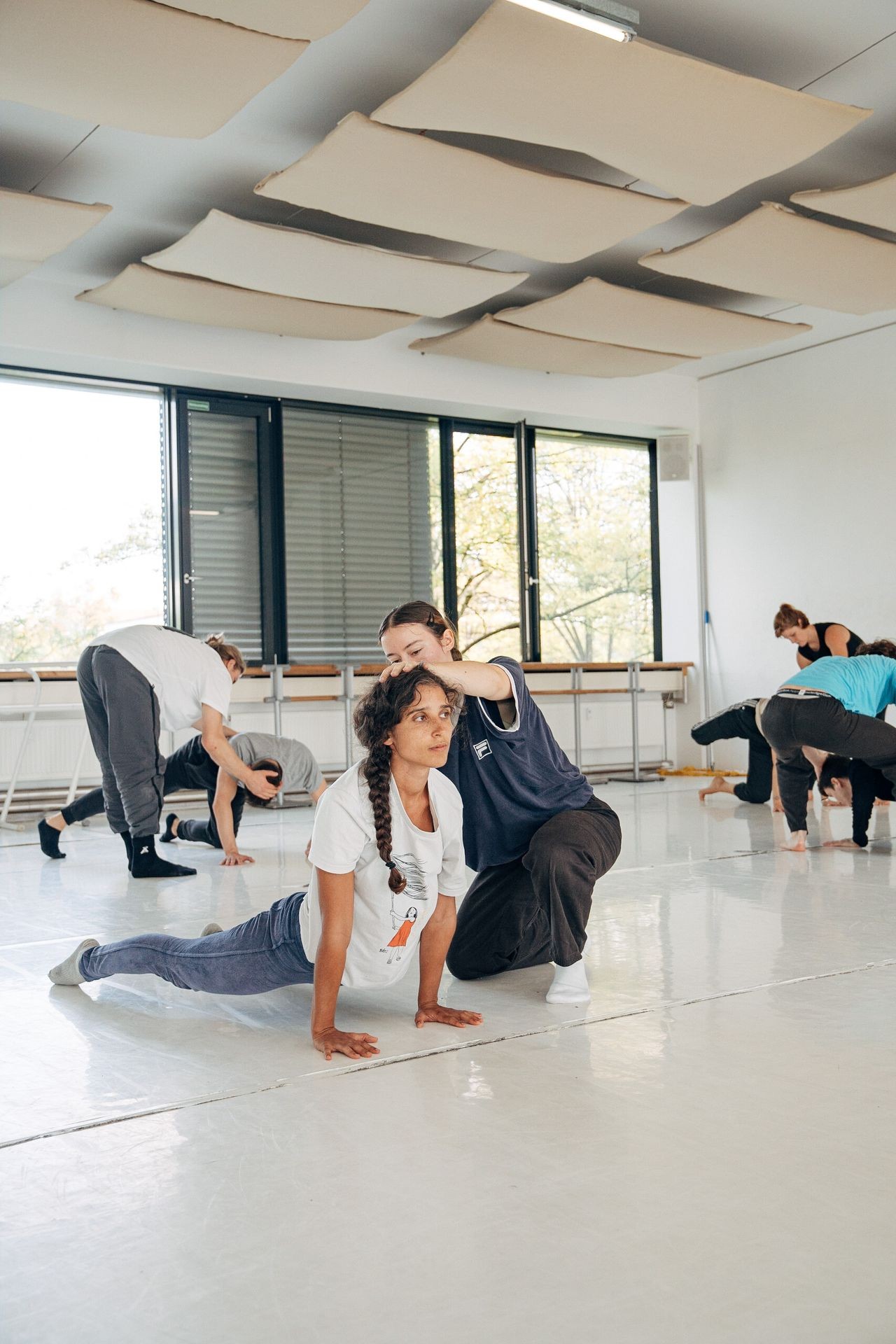 People practicing yoga poses in a bright studio with large windows and a white floor.